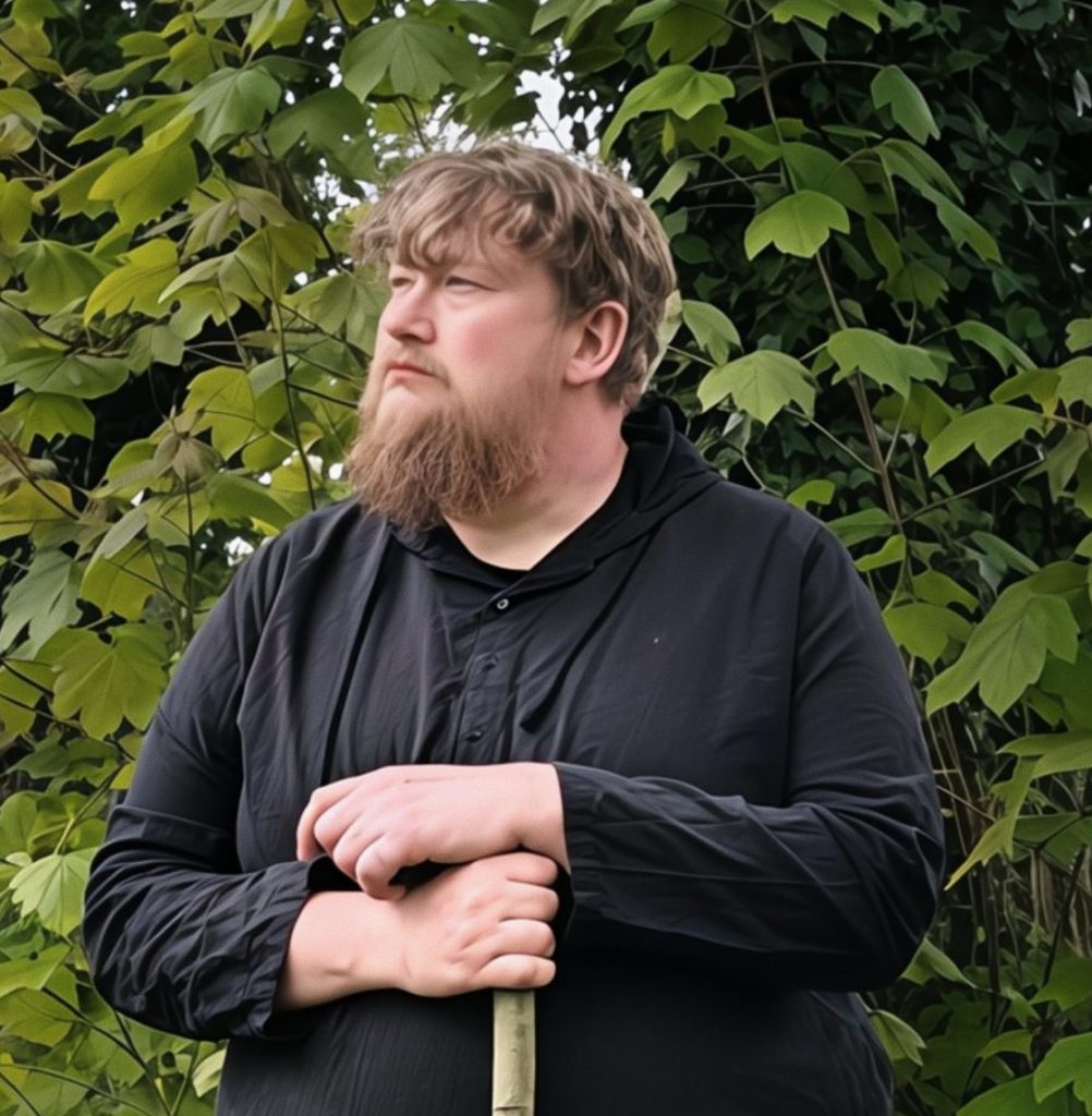 A bearded man in a black robe stands with a staff in a lush garden. He's surrounded by green foliage and yellow flowers, gazing thoughtfully.