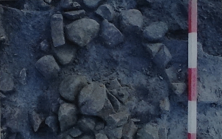 An archaeological excavation of a Mesolithic site with a circular arrangement of rocks on a dusty surface. A red and white measuring stick is placed vertically on the right.
