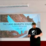 A man stands in front of a projected presentation titled "The Land Beneath Your Feet," with a mural of blue wings on a brick wall background. He wears a "Moon Books" t-shirt, conveying an intellectual tone.