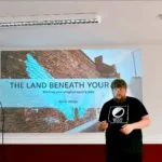 A man stands in front of a projected presentation titled "The Land Beneath Your Feet," with a mural of blue wings on a brick wall background. He wears a "Moon Books" t-shirt, conveying an intellectual tone.