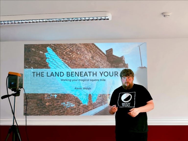 A man stands in front of a projected presentation titled "The Land Beneath Your Feet," with a mural of blue wings on a brick wall background. He wears a "Moon Books" t-shirt, conveying an intellectual tone.