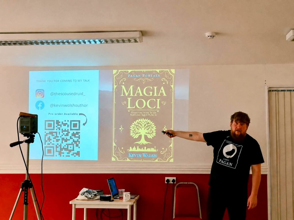 A man presents "Magia Loci" book cover on a screen. He points with a remote, wearing a "Moon Books" T-shirt. QR code and social media handles are displayed.