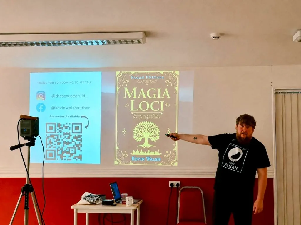 A man presents "Magia Loci" book cover on a screen. He points with a remote, wearing a "Moon Books" T-shirt. QR code and social media handles are displayed.