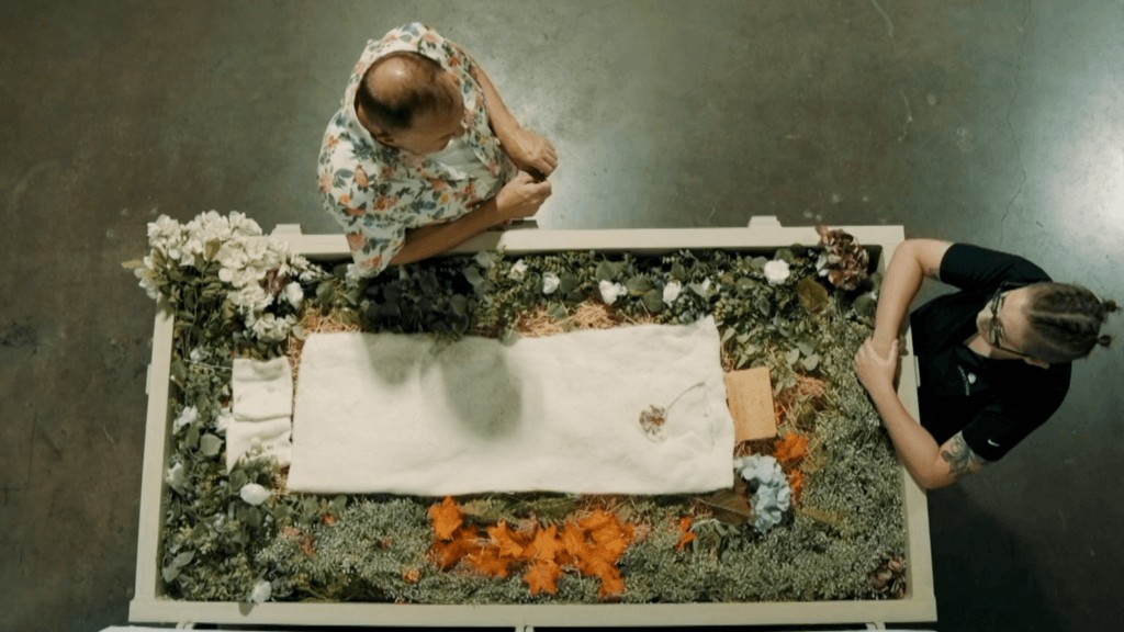 Overhead view of two people beside a flower-adorned terramation vessel. One wears a floral shirt; the other is in black. Tone is calm and reflective.