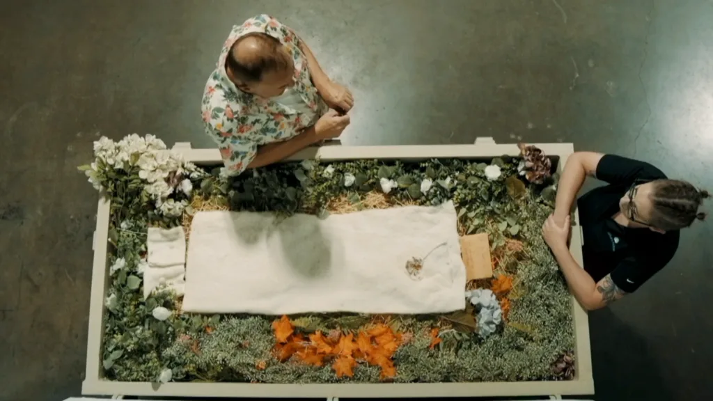 Overhead view of two people beside a flower-adorned terramation vessel. One wears a floral shirt; the other is in black. Tone is calm and reflective.