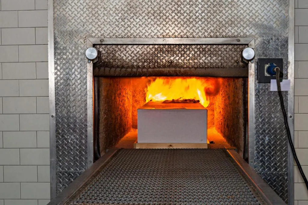 A cardboard box is engulfed in flames inside a cremation chamber, with an industrial, metallic exterior and tiled walls, conveying a somber tone.