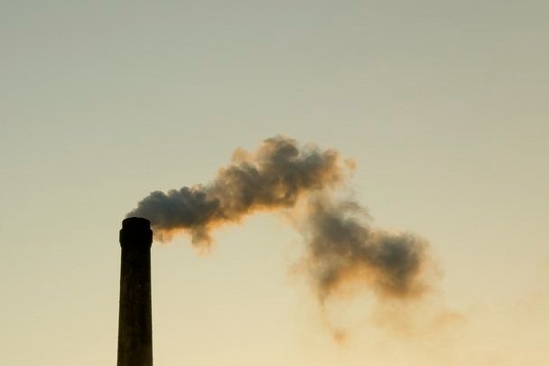 A tall smokestack emits thick, dark smoke against a pale sky, suggesting pollution and environmental impact, with a somber, industrial tone.