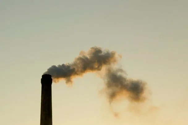 A tall smokestack emits thick, dark smoke against a pale sky, suggesting pollution and environmental impact, with a somber, industrial tone.